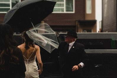 Limo driver holding umbrella for bride as she gets into limo with her gown blowing in the wind